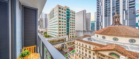 A balcony with a wooden table and chair, overlooking a cityscape with modern and historic buildings, including a church with a dome.