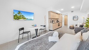 Ein modernes Wohnzimmer mit einer Couch, einem Essbereich mit Tisch und Stühlen, einer Küche mit Holzausstattung und einem großen Fenster mit Blick auf einen tropischen Strand.
