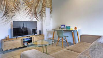 Un salon moderne avec une table basse en verre, un canapé, un téléviseur posé sur un meuble en bois, un bureau avec une chaise et un mur orné d’un motif de feuilles de palmier.