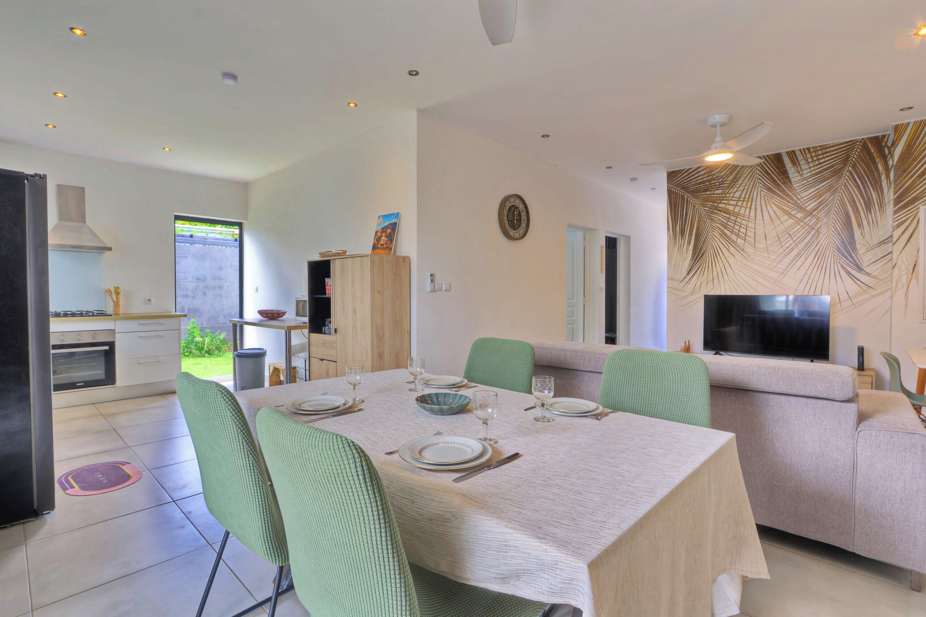 Un espace de salle à manger moderne avec une table dressée pour quatre personnes, des chaises vertes, un téléviseur et une cuisine en arrière-plan.