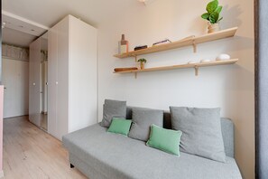 Ein modernes Schlafzimmer mit grauem Sofa, grünen Kissen, Holzbögen mit Büchern und einer Pflanze sowie einem Kleiderschrank.