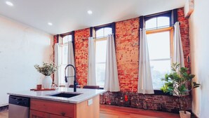 A kitchen with a brick wall, a window with curtains, a sink, and a potted plant. - American Hall Kaufman Suite (Millersburg)