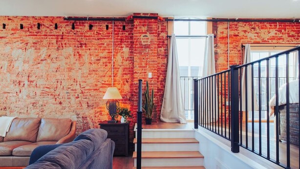 A living room with a brick wall, a sofa, a lamp, and a staircase with a railing. - American Hall Kaufman Suite (Millersburg)