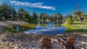 Cena serena à beira de um lago, com duas cadeiras Adirondack de frente para um espelho d'água tranquilo, cercado por árvores e céu claro.