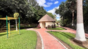 Una cabaña con techo de paja, un área de juegos con juegos de color amarillo y verde, y un sendero de jardín bien cuidado.