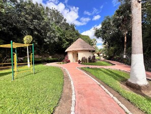A thatched-roof hut, a playground with yellow and green equipment, and a well-maintained garden path. - Cozy Home with Private Pool | 2BD 3BR (Puerto Morelos)