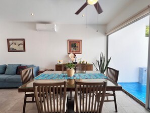 A dining area with a table set for four, a ceiling fan, and a view of an outdoor swimming pool. - Cozy Home with Private Pool | 2BD 3BR (Puerto Morelos)