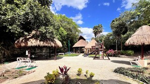 Un parque infantil con tobogán, columpios y una estructura para escalar, rodeado de árboles y cobijados por refugios de paja.
