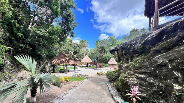 Un sendero conduce a un parque infantil con construcciones de techo de paja, rodeado de exuberante vegetación y un afloramiento rocoso.