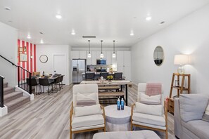 A modern living room with a dining area, a kitchen in the background, and a staircase leading to another level.
