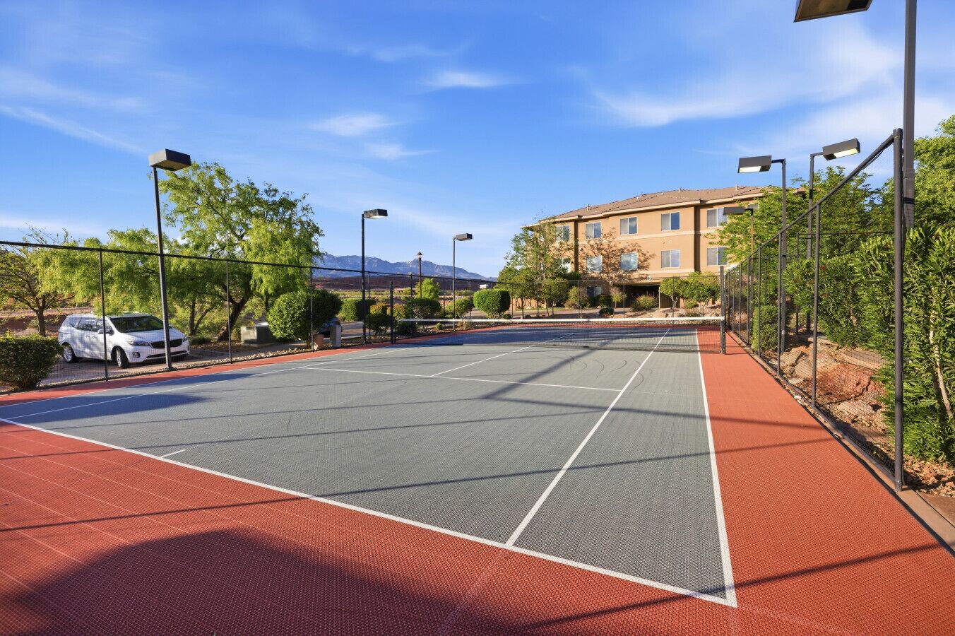 Una cancha de tenis al aire libre con superficie roja, rodeada por una valla, árboles y un edificio al fondo.