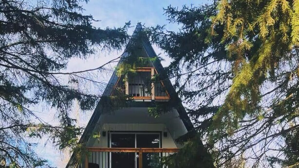 A modern, triangular-shaped house with a glass entrance, wooden deck, and surrounded by trees.