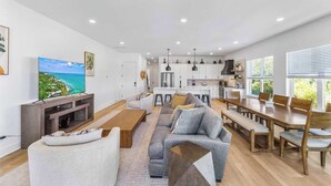 Una sala de estar moderna con sofá, mesa de centro y televisor. Hay una zona de comedor con mesa y sillas. Se ve la cocina al fondo, con armarios y una encimera.