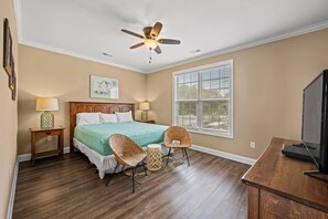 Une chambre à coucher comprenant un lit, deux tables de chevet, un ventilateur de plafond, une fenêtre avec des stores et un téléviseur posé sur un meuble en bois.