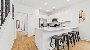 A modern kitchen with white cabinetry, a central island with stools, stainless steel appliances, and a wooden staircase.
