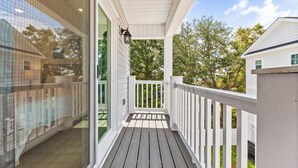 A covered porch with a white railing, a glass door, and a view of trees and a house in the background.