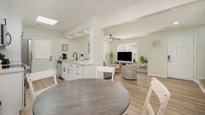 Una cocina moderna con armarios blancos, una mesa de comedor y sillas. En la sala de estar hay un televisor y un ventilador de techo.