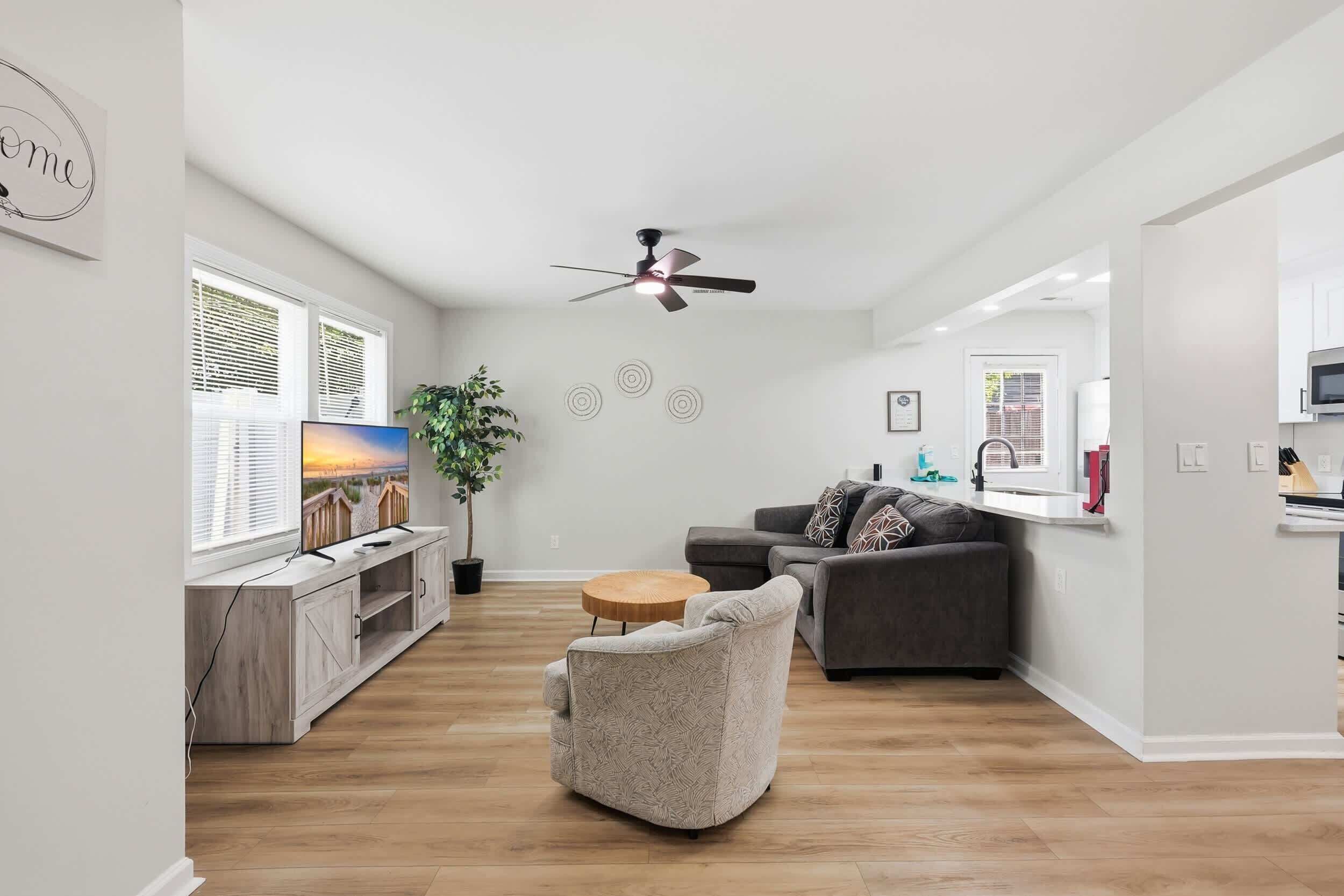 Ein modernes Wohnzimmer mit grauem Sofa, einem Holztisch, einem Fernseher auf einem Holzständer, einem Deckenventilator und einem großen Fenster mit Jalousien.