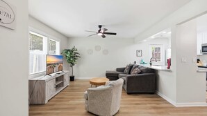 A modern living room with a grey sofa, a wooden coffee table, a TV on a wooden stand, a ceiling fan, and a large window with blinds. - Seabreeze Retreat Modern Home Walk to Beach (Myrtle Beach)