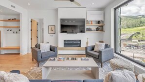 Een moderne woonkamer met een open haard, twee banken, een salontafel en een groot raam met uitzicht op de omgeving.