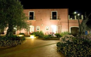 A building with a paved entrance, a small garden with plants and flowers, and a balcony with a railing. - Agriturismo Badiula (Carlentini)
