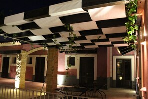 An outdoor seating area with tables and chairs under a black and white checkered awning, surrounded by stone pillars and a building with doors and windows. - Agriturismo Badiula (Carlentini)