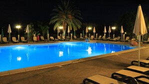 Une piscine entourée de palmiers et dotée d’un éclairage nocturne, avec des chaises longues et des parasols à proximité.