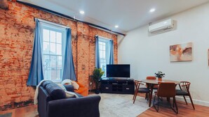 A living room with a brick wall, a sofa, a TV, a dining table with chairs, and a potted plant. - American Hall Young Suite (Millersburg)