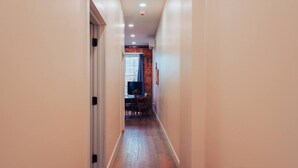 A narrow hallway with a wooden floor, a door on the left, and a room with a brick wall and a window in the background. - American Hall Young Suite (Millersburg)