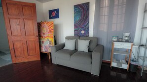Ein Zimmer mit grauem Sofa, Holztür, abstrakten Bildern, einer Kaffeemaschine und einem Metallregal.