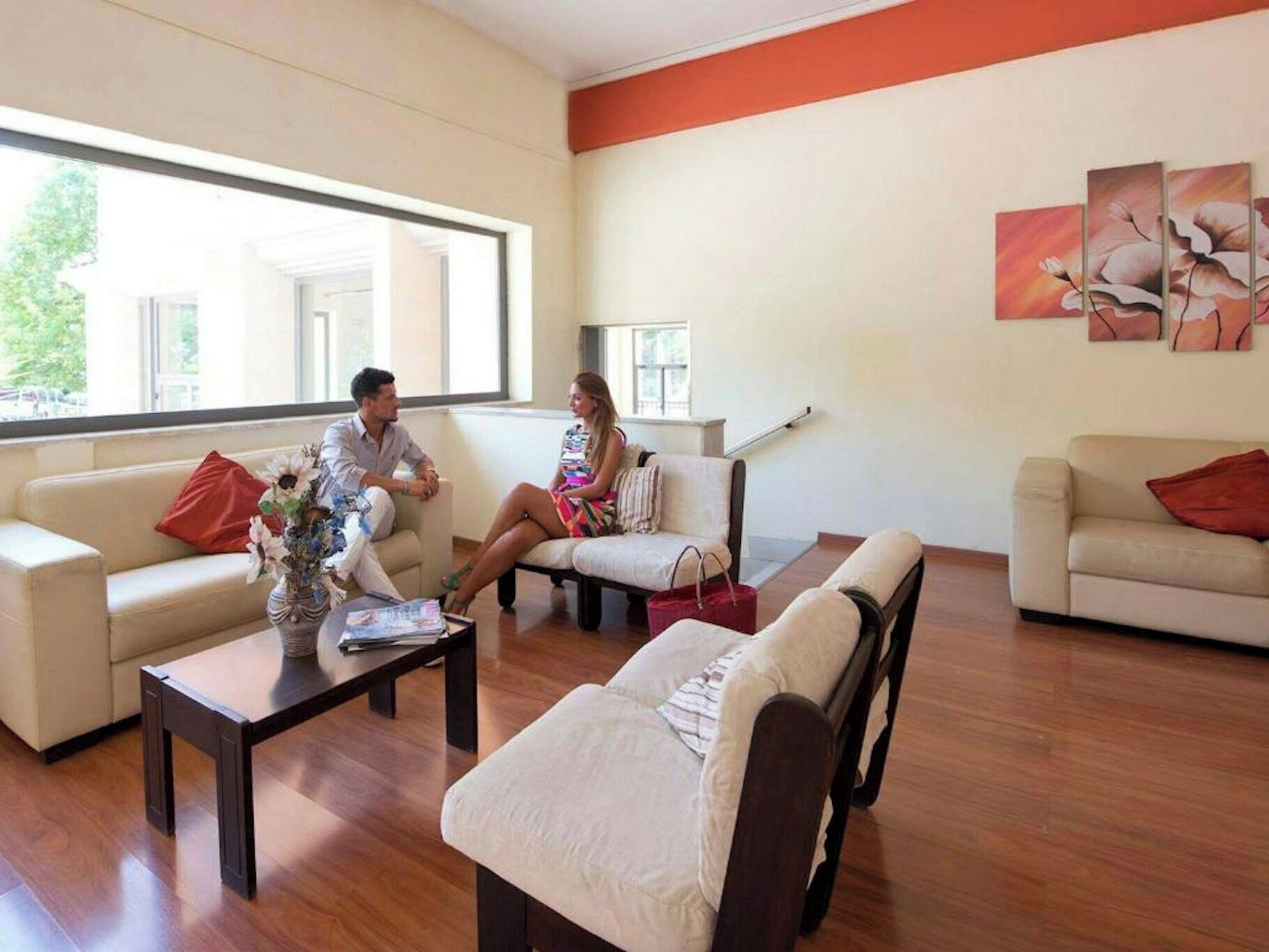 A spacious living room with a large window, a coffee table with a vase, and a sofa with a red pillow.