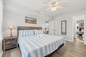 A bedroom with a bed, bedside tables, a ceiling fan, a painting, and a door leading to another room. - Wavecrest Grand with Pool Great for Groups (Surfside Beach)