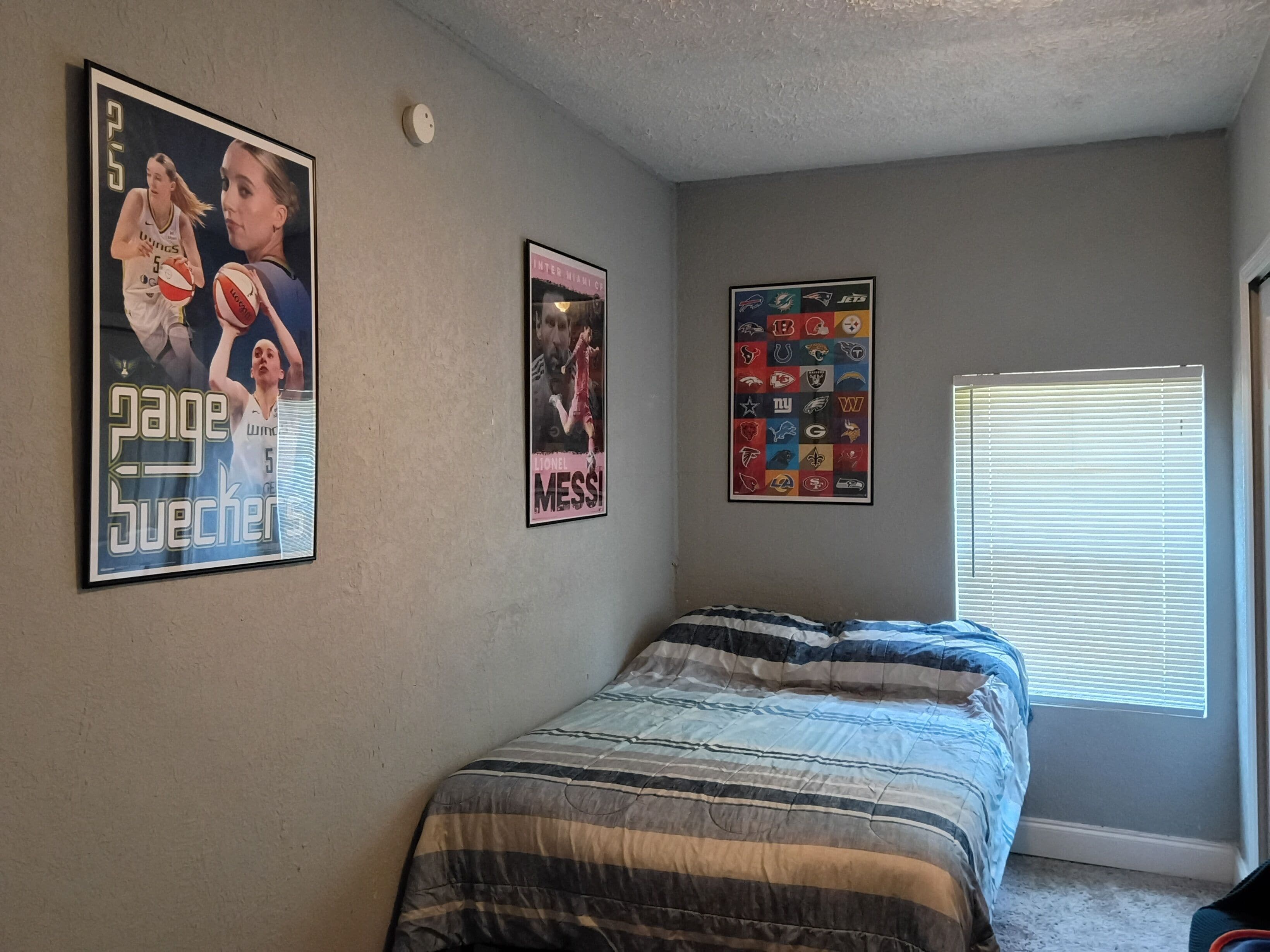 A bedroom with a bed, a poster of Paige Bueker, a poster of Lionel Messi, and a poster of NFL team logos.