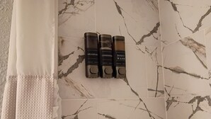 A bathroom with a marble floor, a towel rack with three bottles of shampoo, conditioner, and body wash, and a white shower curtain. - THE JAX HIDEOUT 
DT studio w/shaded patio (Jacksonville)