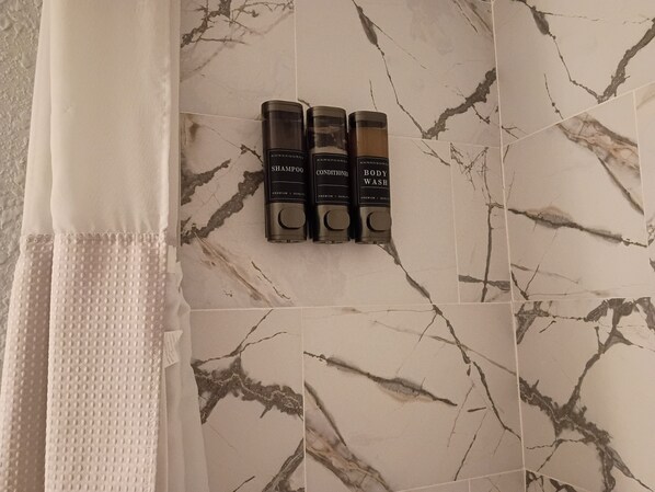A bathroom with a marble floor, a towel rack with three bottles of shampoo, conditioner, and body wash, and a white shower curtain.