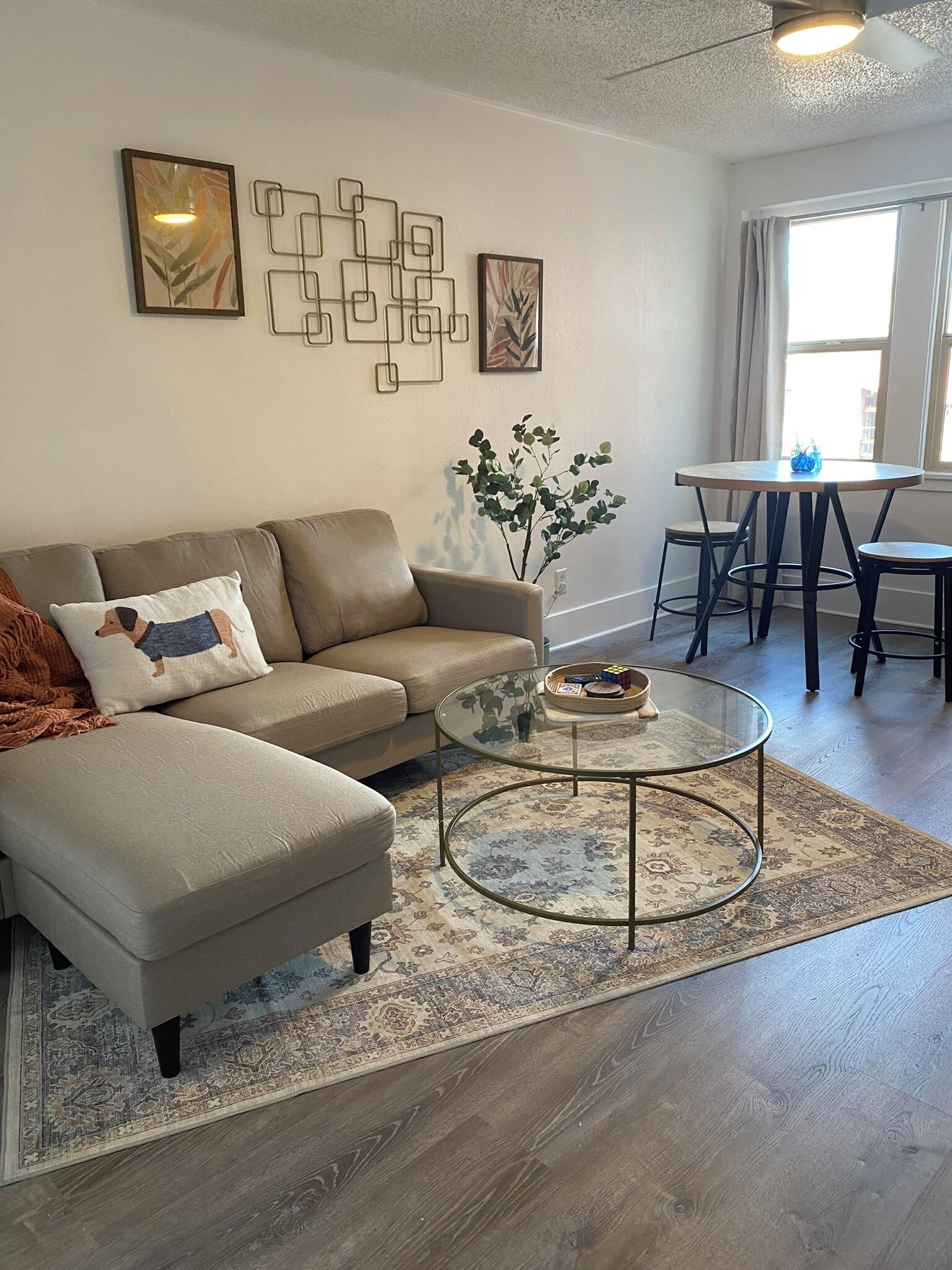 A living room with a sectional sofa, a glass coffee table, a patterned rug, and a dining area with a table and chairs.