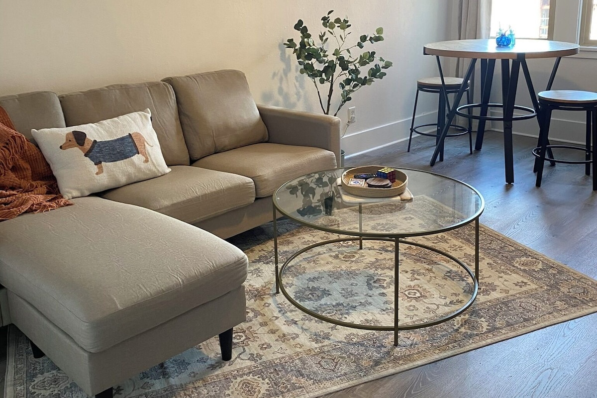 Un salon comprenant un canapé d’angle, une table basse en verre, un tapis à motifs et un coin repas avec une table et des chaises.