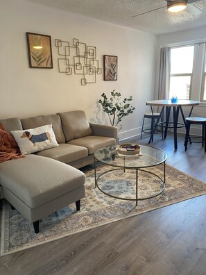 A living room with a sectional sofa, a glass coffee table, a patterned rug, and a dining area with a table and chairs. - The Historic George Hotel (Klamath Falls)