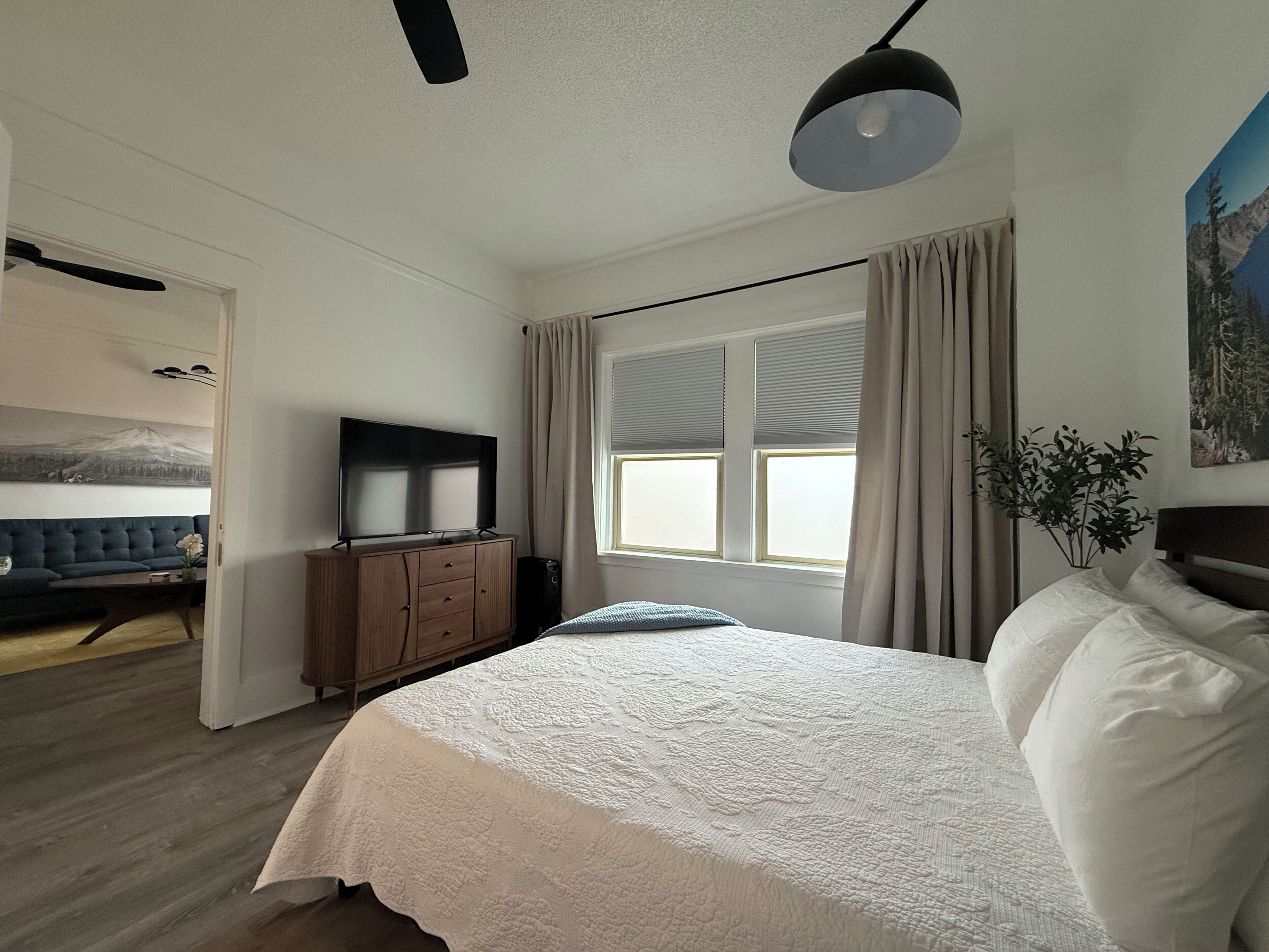A bedroom with a bed, a TV on a wooden cabinet, a window with curtains, and a painting on the wall.