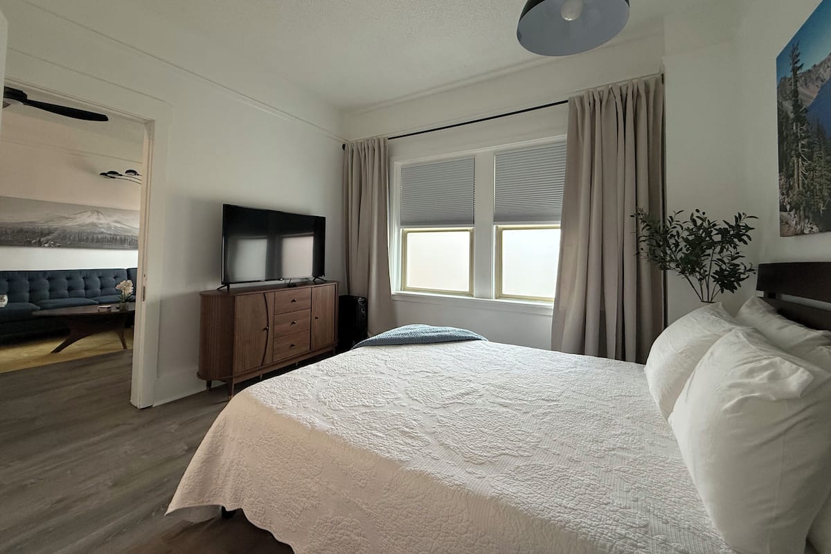 A bedroom with a bed, a TV on a wooden cabinet, a window with curtains, and a painting on the wall.