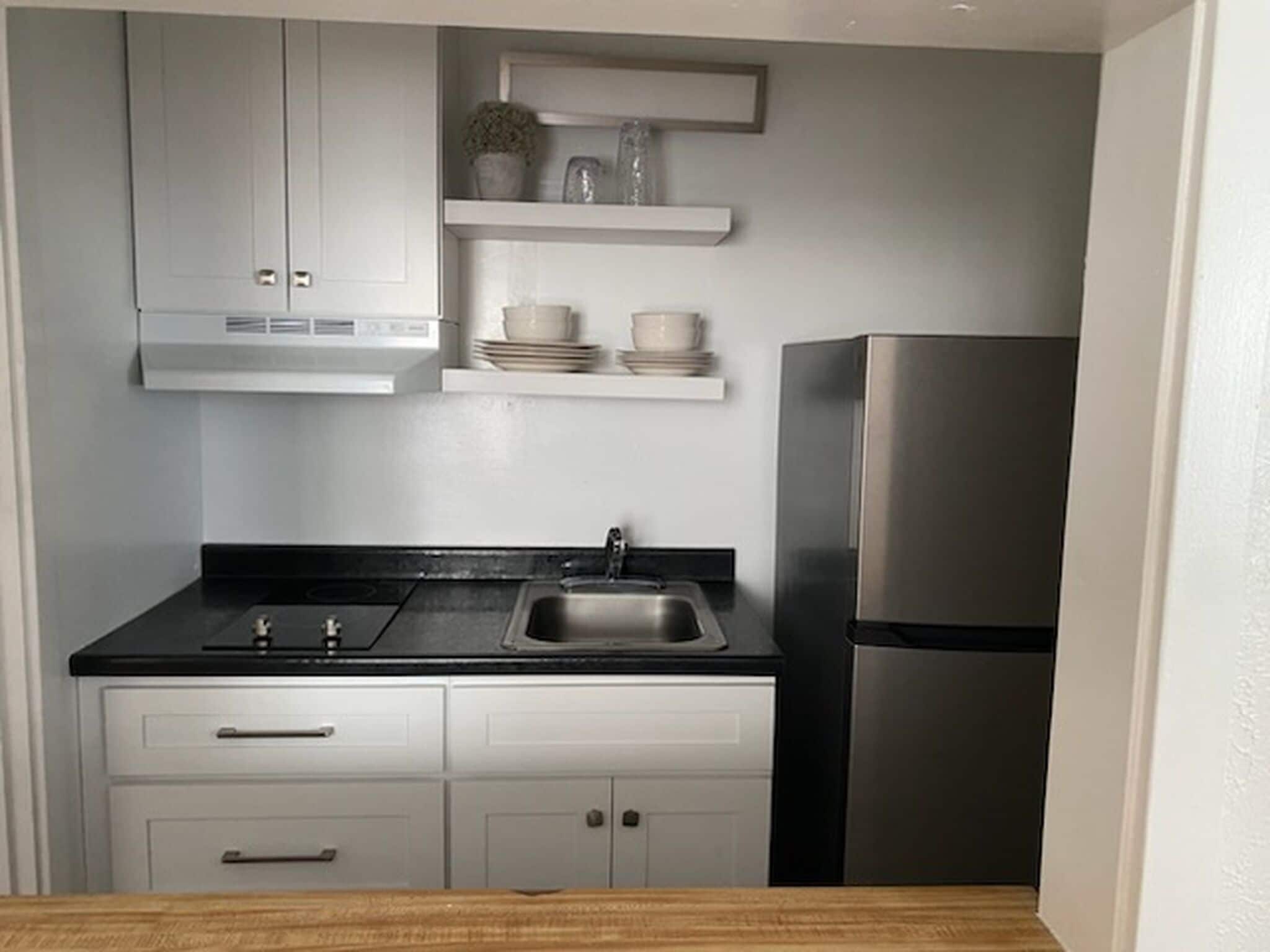 A compact kitchen with a stainless steel refrigerator, a black countertop, a sink, and white cabinetry.