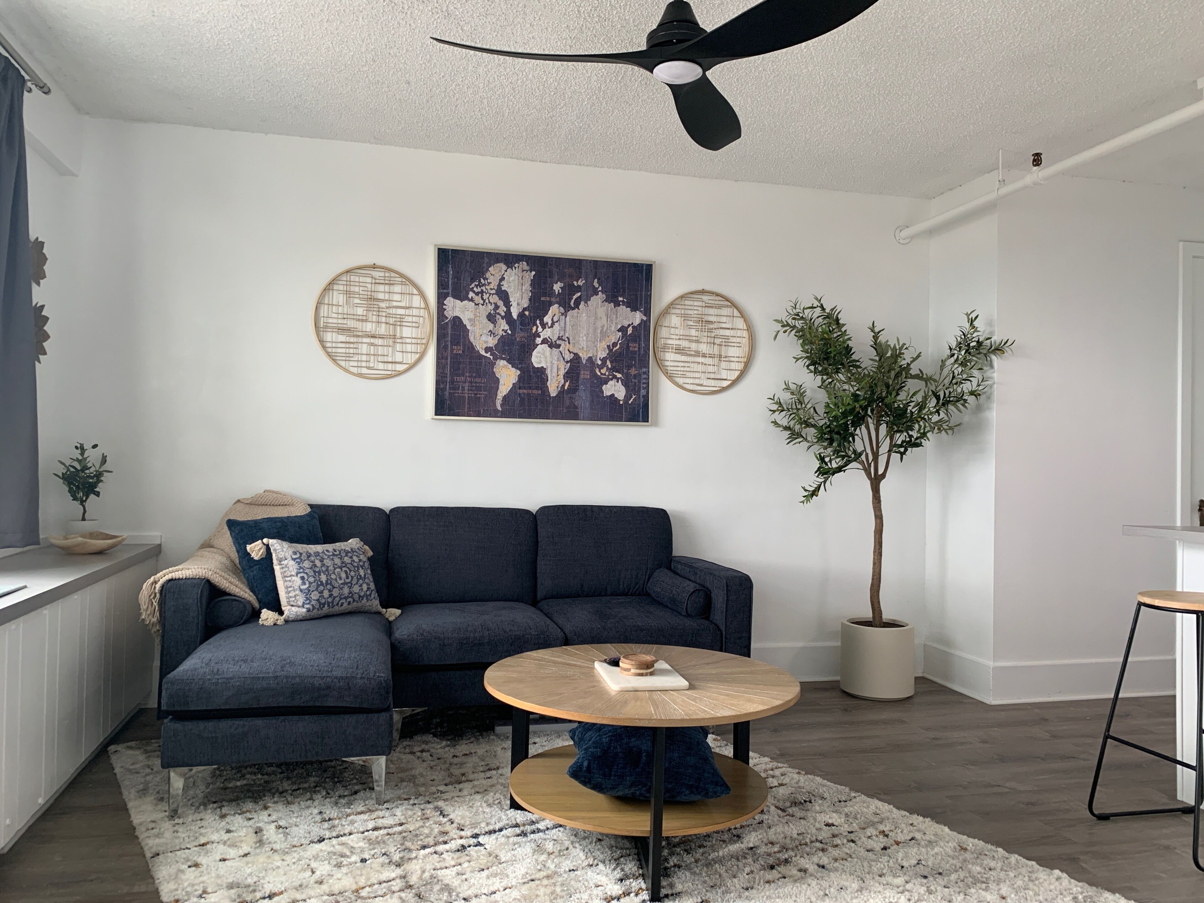 A modern living room with a dark blue sectional sofa, a round wooden coffee table, a world map artwork, and a potted tree.