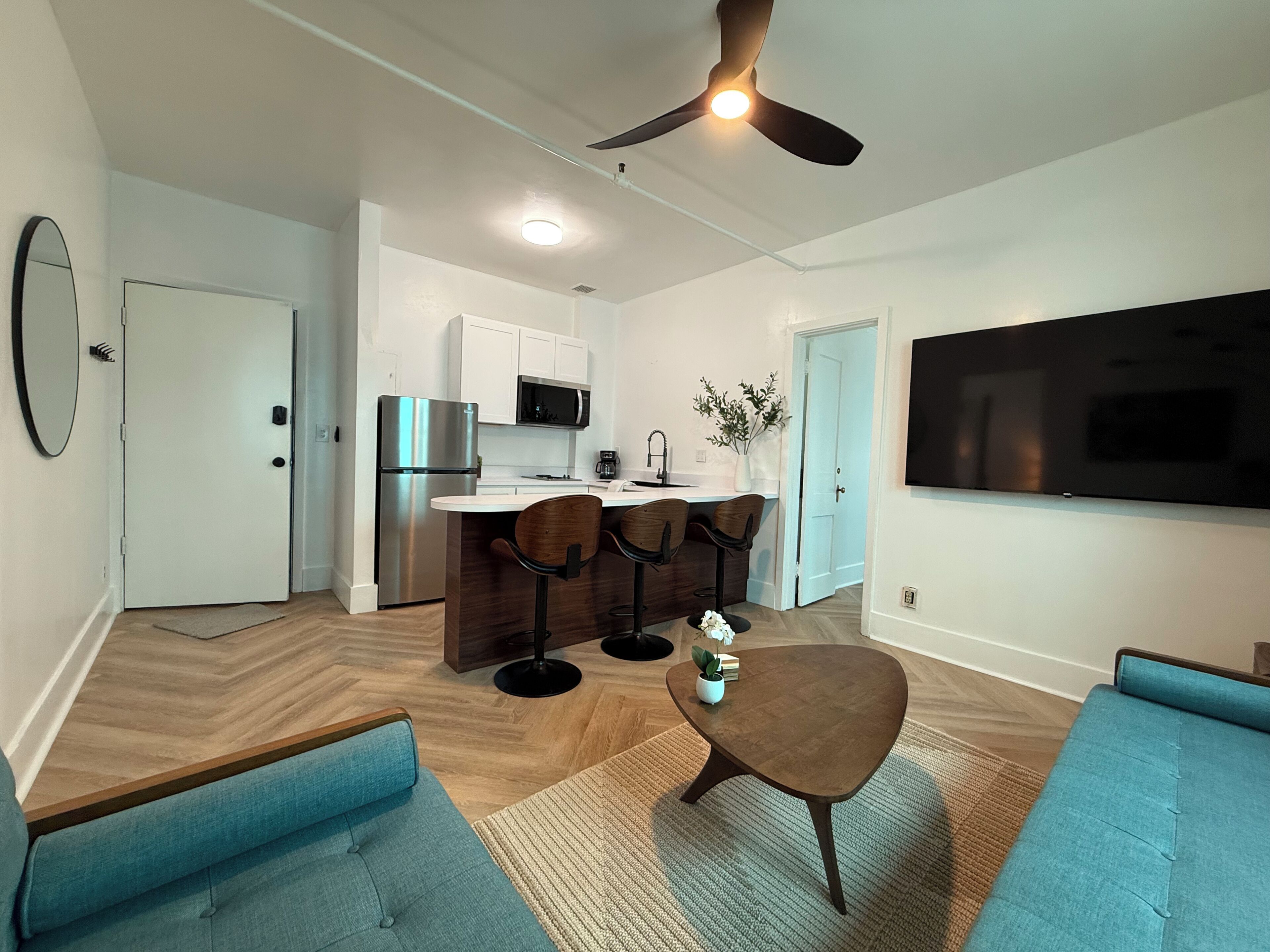 A modern living room with a sofa, a coffee table, a TV, and a kitchen area with a fridge and bar stools.