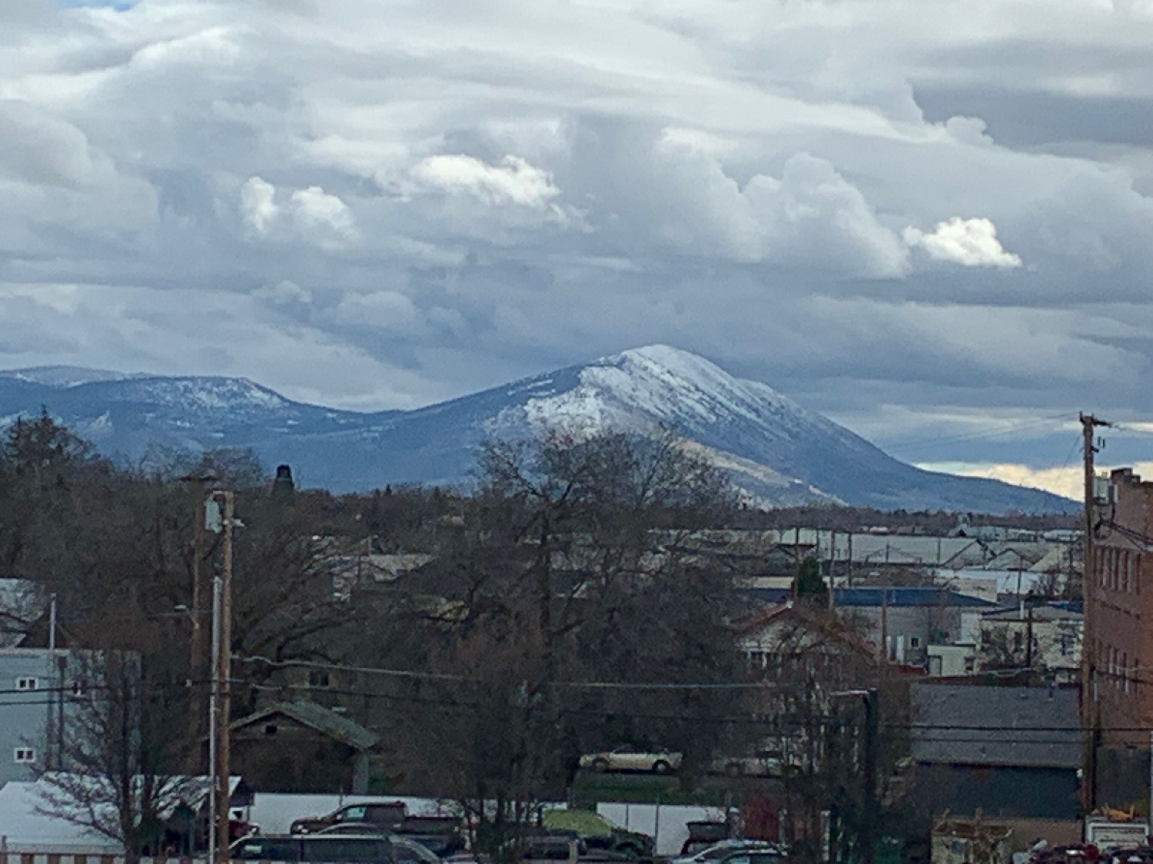 A mountain with snow-capped peaks in the background, a parking lot with several cars, and buildings with flat roofs in the foreground.