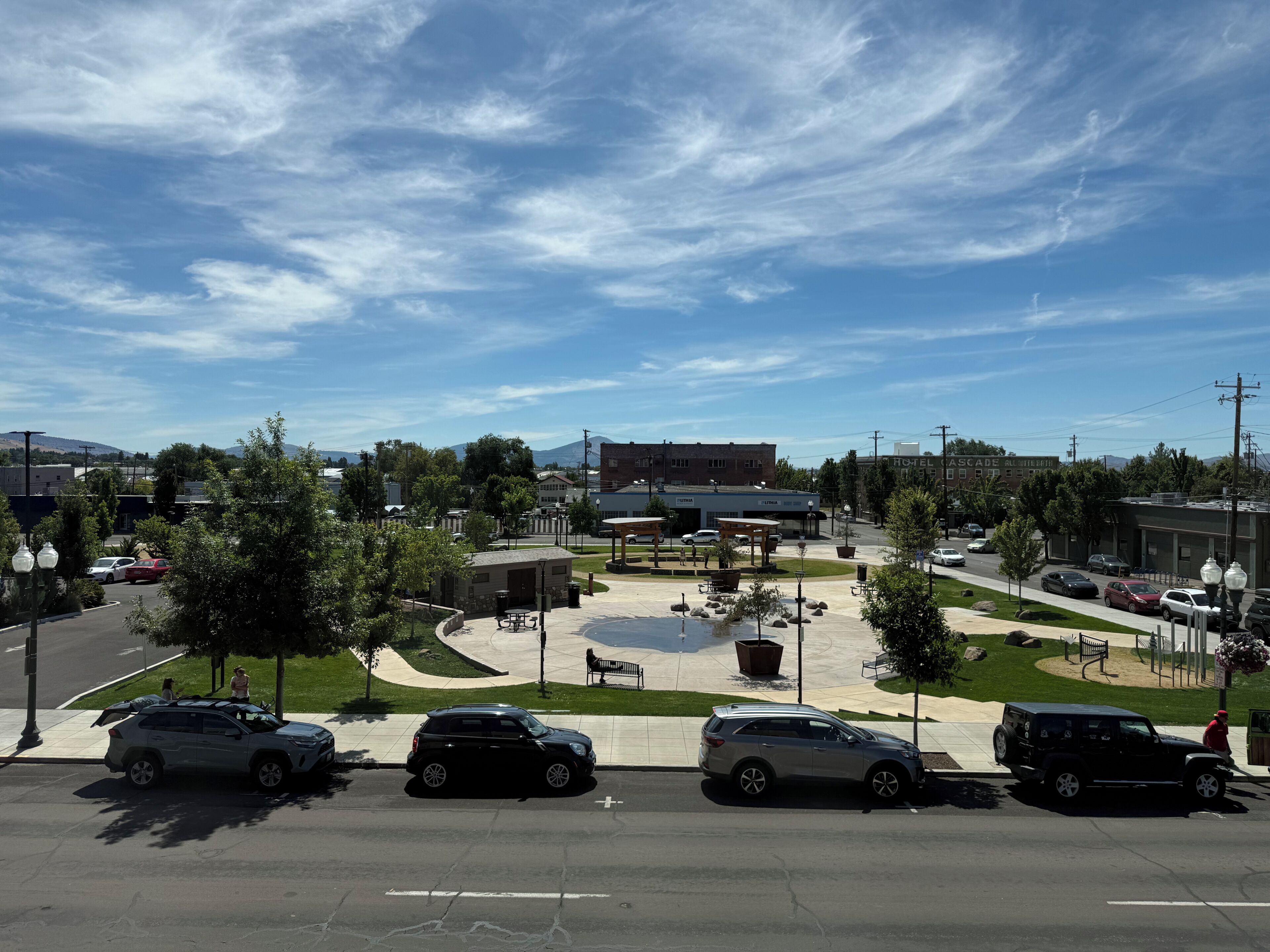 A parking lot with multiple cars, a central plaza with benches, and buildings in the background.