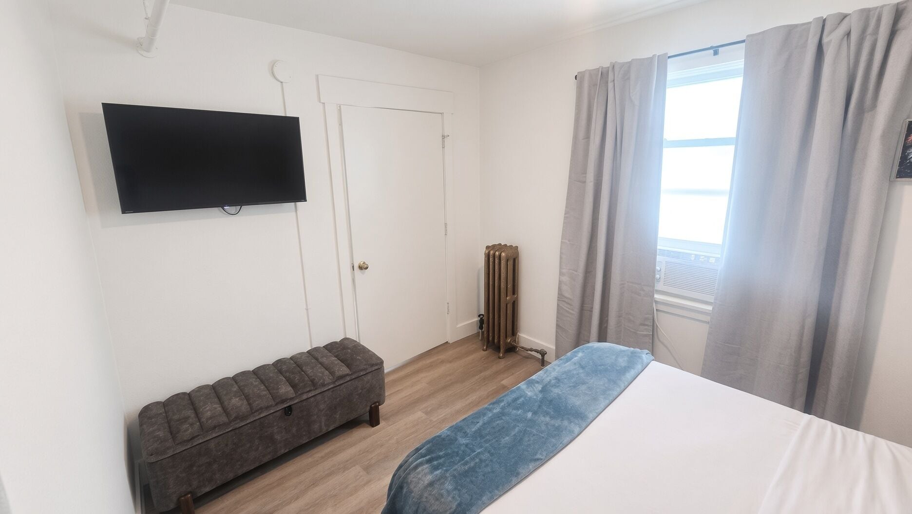 Une chambre à coucher avec un lit, un banc, un radiateur, une télévision et une fenêtre avec des rideaux.