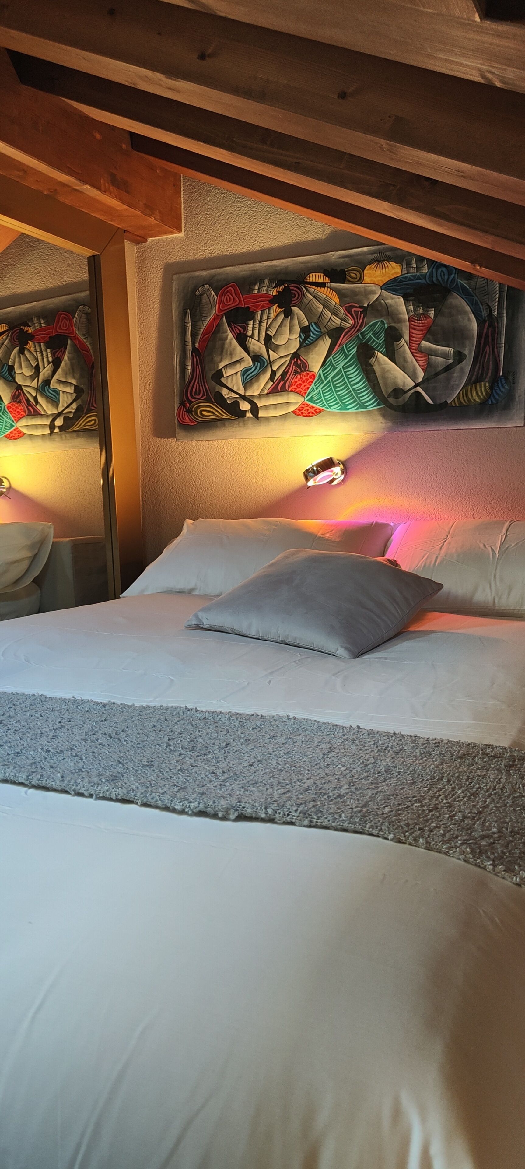 Ein Bett mit weißer Bettwäsche, einem grauen Kopfkissen und einer gemusterten Decke. Darüber hängen zwei Bilder an der Wand.
