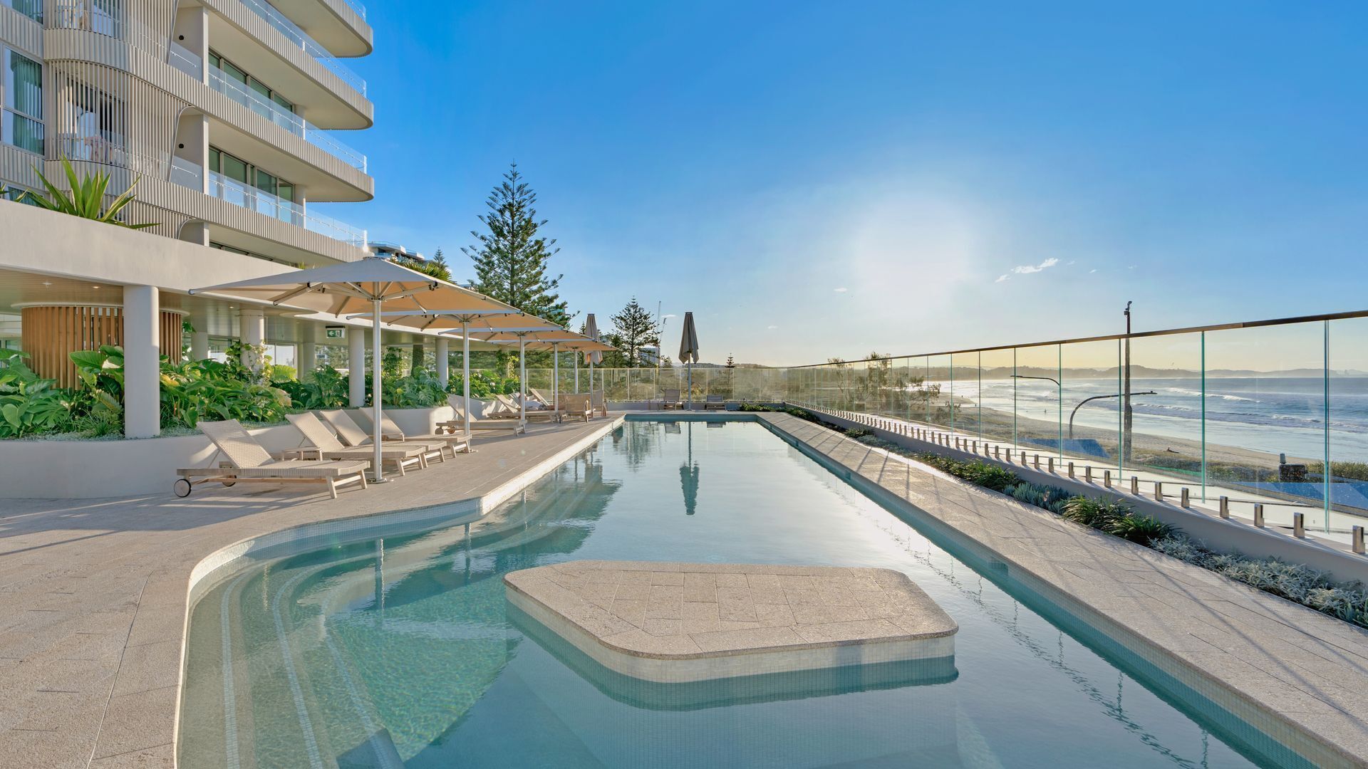 Une piscine moderne d’hôtel, dotée d’une balustrade en verre, de chaises longues et offrant une vue sur l’océan.