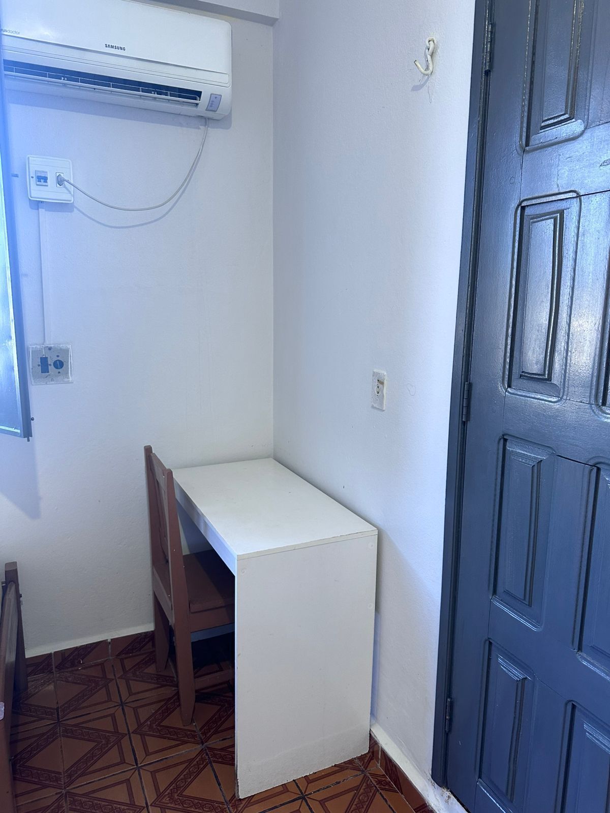 A small room with a white desk, a chair, a blue door, and an air conditioning unit mounted on the wall.