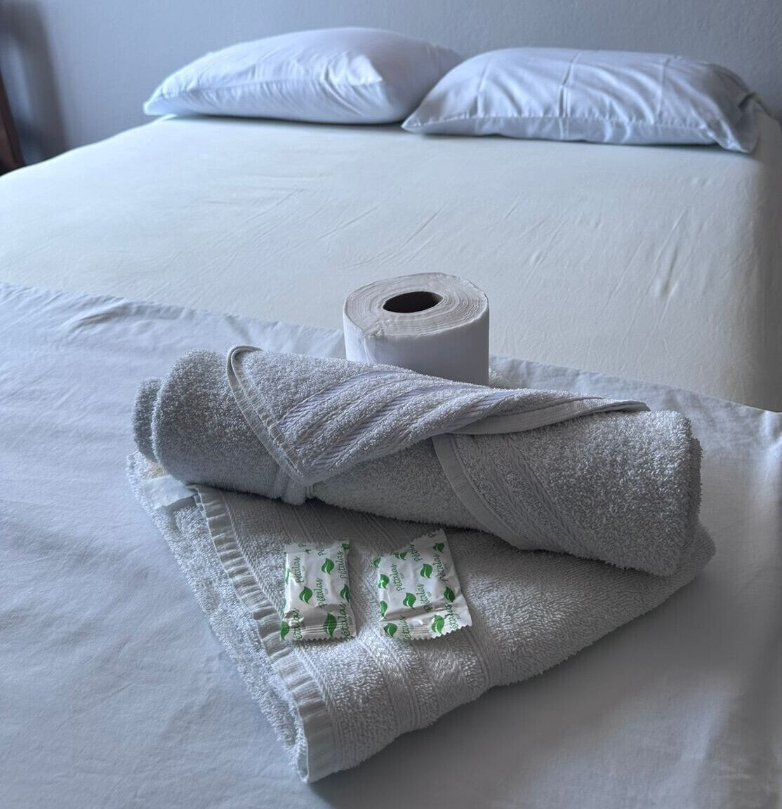 A neatly made bed with white linens, a rolled-up towel, and a pair of slippers.